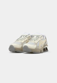 Beige athletic shoes with mesh panels, contrasting gray soles, and beige accents, featuring a lace-up design and logo on the tongue.