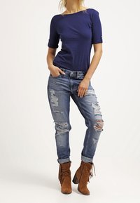 Marinblå t-shirt med uppvikt ärm, ihop med slitna blå jeans med hål och bruna mocka ankelstövlar med fransdetaljer.