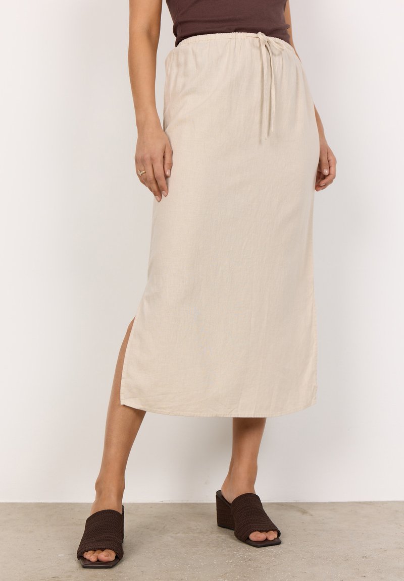 Beige, knielanger Rock mit elastischem Bund und Zugband, ausgestattet mit seitlichen Schlitzen. Kombiniert mit dunkelbraunen, strukturierten Blockabsatzsandalen.