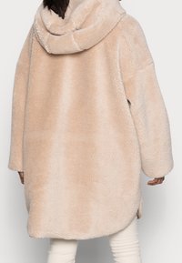 Oversized beige fleecetrøje med hætte, der har en blød tekstur og afrundet kant, designet til komfort og varme.