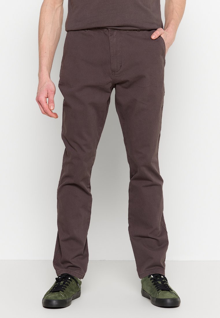 Pantalon en coton marron pour homme avec une coupe droite, doté d'une braguette à boutons et d'un design à cinq poches, associé à des baskets noires et vertes.
