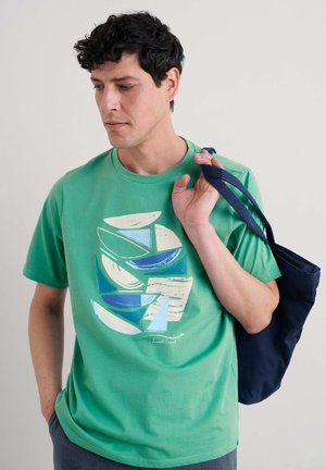 Camiseta de algodón verde con patrones geométricos abstractos y multicolores. El modelo lleva un bolso de mano azul marino colgado de un hombro.