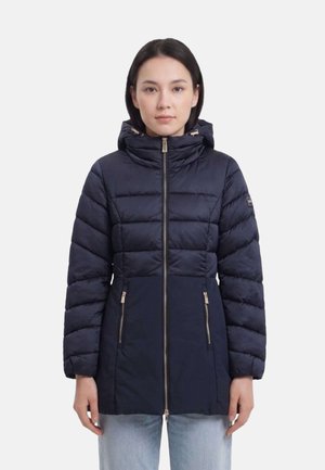 Donna che indossa una giacca invernale trapuntata blu navy con cappuccio, cerniera frontale e tasche con cerniera, in piedi davanti a uno sfondo semplice.