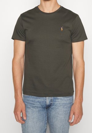Mężczyzna noszący gładki, ciemnozielony t-shirt z krótkim rękawem z małym wyszywanym logo na lewej piersi oraz jasnoniebieskie dżinsy.