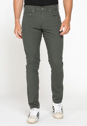 Uomo che indossa pantaloni slim fit verde scuro e sneakers bianche con strisce nere in piedi davanti a uno sfondo semplice.