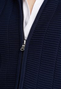 Cárdigan de punto con cremallera en color azul marino, con un brillo plateado en la cremallera. Debajo, se puede ver parcialmente una camisa blanca con cuello.