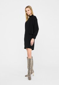 Abito lungo nero con maniche a sbuffo e dettaglio in cappotto fluente, abbinato a stivali alti in pelle di serpente a motivi e una silhouette slim.