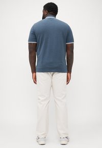 Man met tatoeages in een blauw overhemd met korte mouwen, ivoorkleurige broek en sneakers, staand met de rug naar de camera tegen een effen achtergrond.