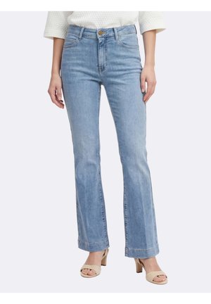 Vrouw die lichtblauwe jeans met wijde pijpen en hoge taille draagt, gecombineerd met beige open-toe sandalen met hak, staand tegen een effen achtergrond.