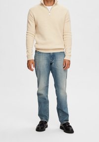 Béžový pletený svetr s límcem na čtvrtý zip, kombinovaný s světle modrými džíny a černými šněrovacími botami. Plochá textura, volný střih.