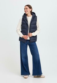 Une femme souriante se tient dans une tenue décontractée avec un gilet matelassé bleu marine, un pull crème et un jean à jambes larges, sur un fond clair.
