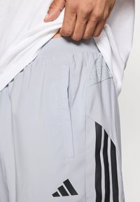 Pantalones cortos deportivos gris claro con rayas negras, un bolsillo lateral y detalles perforados cerca de la cintura. Presentan un logo de adidas.