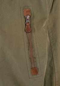 Khaki jacket in durable fabric featuring a vertical zipper with a brown leather pull tab and ribbed accent near the opening.
