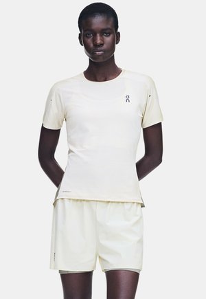 Personne à la peau foncée portant une chemise beige clair à manches courtes et un short assorti, debout avec les mains derrière le dos devant un fond uni.