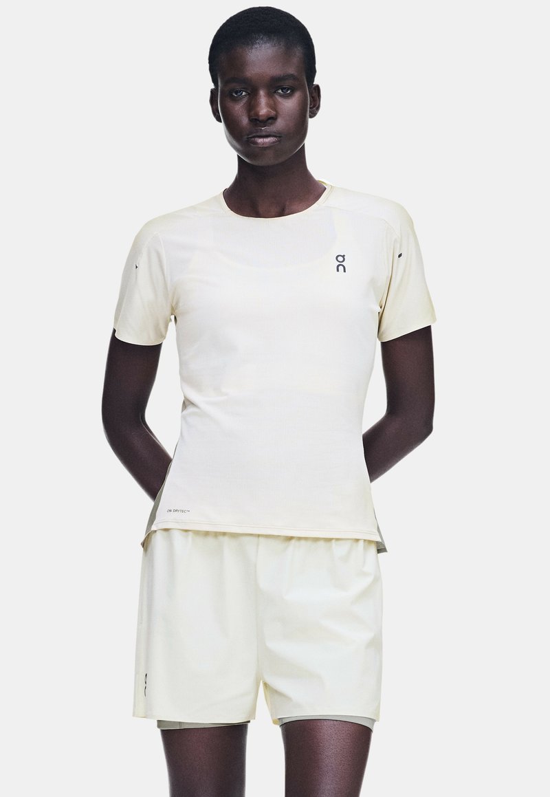 Personne à la peau foncée portant une chemise beige clair à manches courtes et un short assorti, debout avec les mains derrière le dos devant un fond uni.