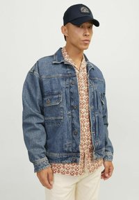 Chaqueta de mezclilla en azul oscuro, con dos bolsillos en el pecho y cierre de botones. Debajo, una camisa de manga corta con un patrón de diseños geométricos.