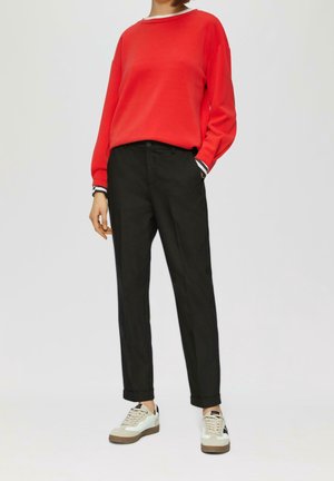 Persona con una camiseta roja de manga larga, pantalones negros con puños y zapatillas blancas con suelas beige sobre un fondo liso.