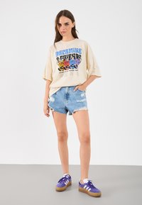 T-shirt oversized creme com uma gráfica de cartoon colorida, combinada com calções de ganga azul desgastados e ténis roxos com riscas brancas.
