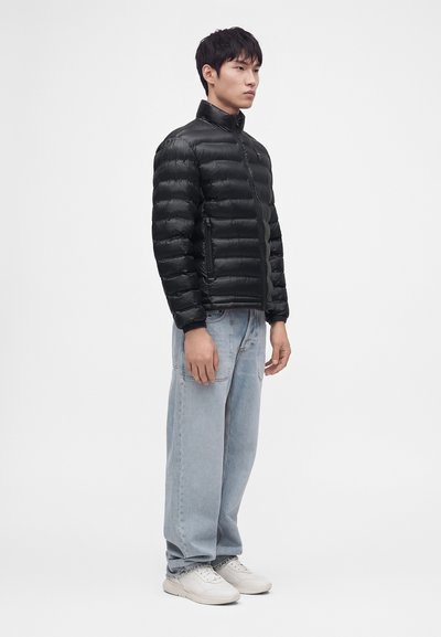 Homme debout, tourné vers la droite, portant une doudoune noire, un jean bleu clair ample et des baskets blanches sur fond blanc.