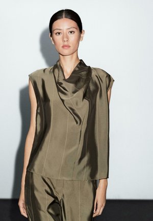 Femme aux cheveux bruns, portant un top sans manches en satin olive vert et un pantalon assorti, se tenant devant un fond clair.