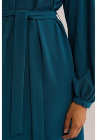 Teal färgad klänning i tyg med knytbart midjebälte, långa ärmar och en mjuk textur med rynkade manschetter. Nära bild av ärm- och bältesdetaljer.