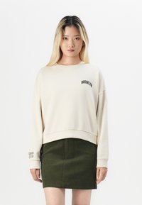 Sweatshirt crème avec "BROOKLYN" brodé, associé à une jupe en velours côtelé vert foncé. Coupe décontractée et manches longues.