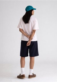 Personne debout, dos à la caméra, portant une casquette bleu sarcelle, une chemise rose clair, un short noir, des chaussettes blanches et des chaussures marron sur fond uni.