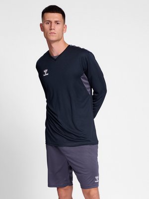 Mann trägt schwarzen langärmeligen Sportshirt und graue Sportshorts, steht mit den Händen hinter dem Rücken vor weißem Hintergrund.