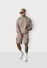 Sweatshirt kaki avec des poignets côtelés, associé à un short cargo assorti. Comprend un col rond, des poches latérales et des baskets blanches.
