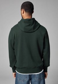 Dark green hooded sweatshirt made from soft fabric, features a kangaroo pocket, ribbed cuffs, and a relaxed fit, worn over blue jeans.