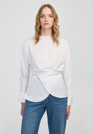 Jonge vrouw met golvend blond haar, gekleed in een witte blouse met lange mouwen en een wikkelontwerp, en blauwe jeans, staande tegen een effen achtergrond.