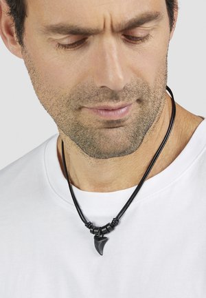 Mann mit Stoppeln, der ein weißes Hemd und eine schwarze Halskette mit einem Haifischzahnanhänger trägt und nach unten schaut.