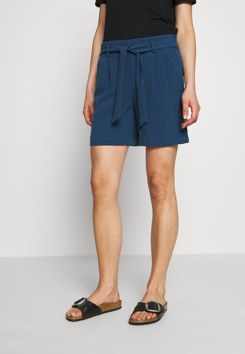 Modré šortky s páskem v bocích a dvěma předními kapsami, vyrobené z hladkého materiálu, ve spojení s černými sandály na klínku se stylovou kovovou sponou.