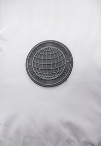 Patch de cauciuc negru cu un design al globului, text embosat "CLOSURE LONDON", pe un fundal de țesătură albă netedă. Detalii cu contrast ridicat.