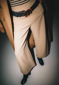 Pantaloni beige a gamba larga con una texture liscia, dotati di una cintura di pelle scura ornata da dettagli decorativi colorati. Stivaletti neri visibili.