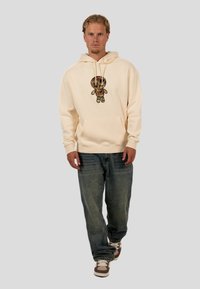 Cream hoodie featuring a colorful graphic design of a doll on the front, paired with light blue jeans and brown sneakers.