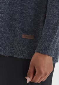 Detail ruky oblečené v tmavě modrém pleteném svetru s hnědou nášivkou "oxmo" nad černými kalhotami.