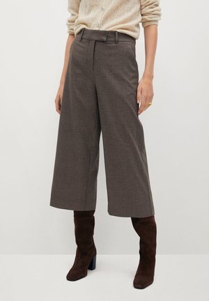 Osoba ubrana w brązowe spodnie w kratę o szerokich nogawkach i wysokim stanie, zestawione z ciemnobrązowymi zamszowymi botkami na obcasie oraz kremowym, teksturowanym swetrem.