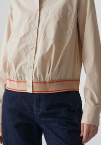 Beige overhemd met knopen en een borstzak, elastische zoom met tan en rode strepen, gecombineerd met donkere denim spijkerbroek, gladde stofstructuur.