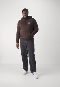 Brun hettegenser med lomme foran og grafisk design på brystet, kombinert med mørkejeans og hvite sneakers, stående på en nøytral bakgrunn.
