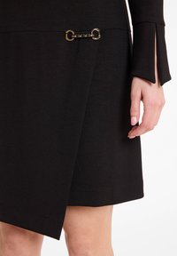 Robe noire avec un design croisé, manches longues avec fentes et une chaîne dorée accentuant la taille. Réalisée dans un tissu lisse et ajusté.