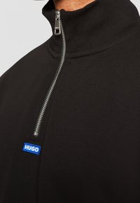 Pull noir à col haut avec demi-fermeture éclair argentée et petit patch rectangulaire bleu avec logo HUGO sous la fermeture éclair sur la poitrine.