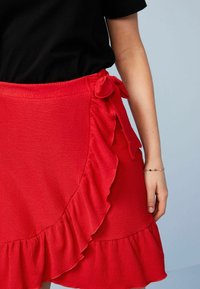 Jupe portefeuille rouge avec ourlet à volants et détail noué sur le côté. Confectionnée en tissu texturé. Associée à un haut noir. Présente un design décontracté et féminin.