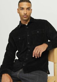 Black corduroy shirt with a button-up front, two chest pockets, and a collar. Worn over dark denim jeans, against a light background.