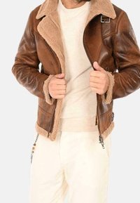 Mężczyzna w brązowej kurtce ze skóry z kożuszkiem, noszący pod nią beżowy sweter i kremowe spodnie, trzymający kurtkę rozpiętą na wysokości klatki piersiowej.