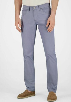 Mann trägt hellblaue Slim-Fit-Hosen und braune Wildleder-Slipper, steht vor einem schlichten weißen Hintergrund.