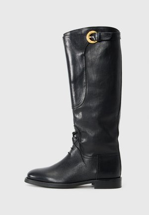 Bota de cuero negro, hasta la rodilla, con punta redonda, parte frontal con cordones, detalle lateral y una hebilla dorada en la parte superior. Textura suave.