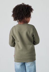 Felpa verde in cotone con polsini e orlo a costine, con dettaglio sulla parte posteriore. Abbinata a jeans denim azzurri per un look casual.