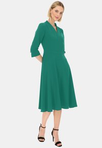 Turkusowa sukienka z dopasowanym korpusem i rozkloszowanym dołem, rękawami do trzech ćwiartek, dekoltem w kształcie litery V oraz subtelnymi guzikami przy rękawach. Czarnie szpilki.