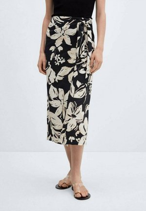 Jupe portefeuille noire avec un motif floral blanc, en tissu léger, longueur mi-mollet, nouée à la taille, associée à des sandales transparentes.